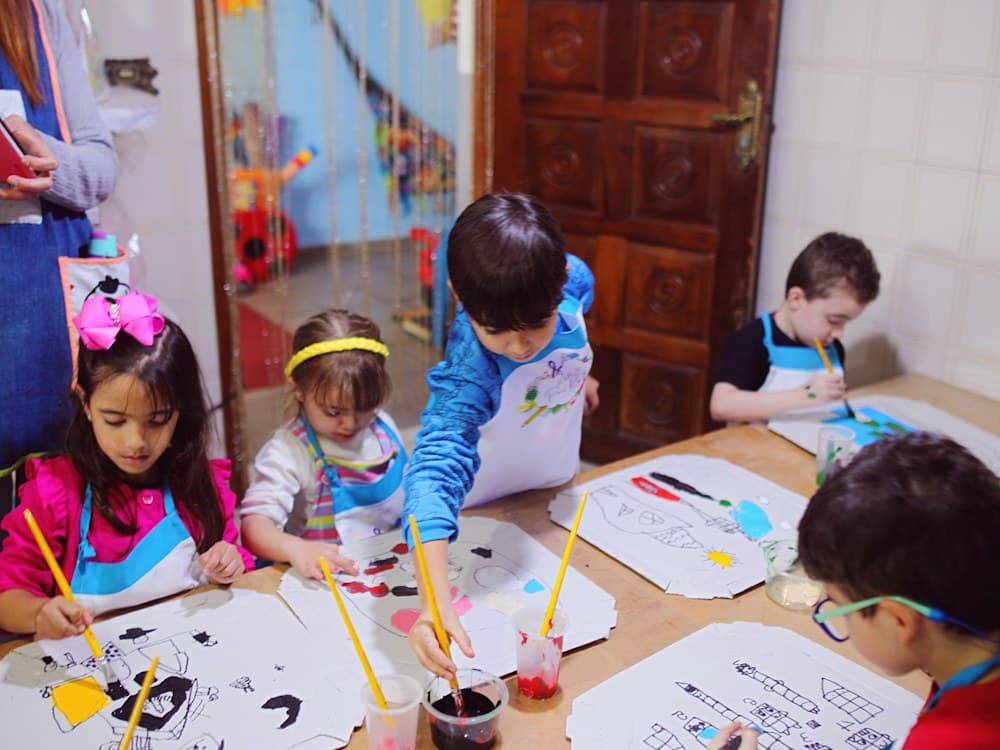 Niñas y niños dibujando y pintando alrededor de la mesa larga de talleres